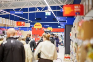 Shoppers walking through a busy supermarket aisle with yellow sale signs and blue overhead category banners.