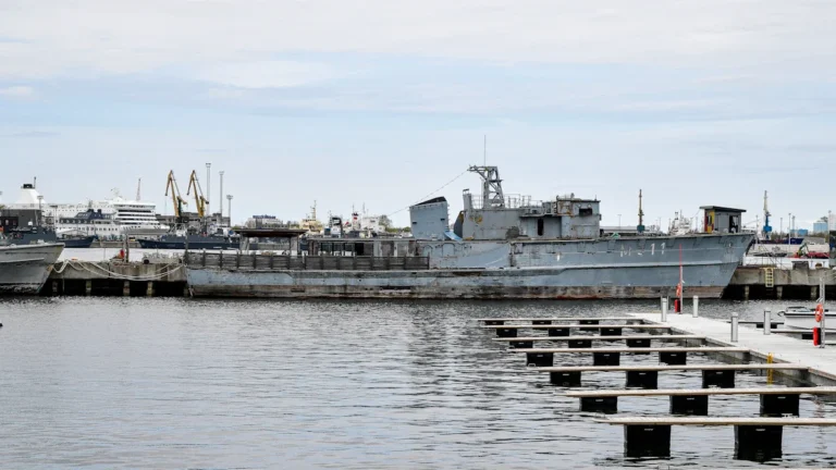 A gray naval ship docked at a harbor pier, illustrating dual-use military and commercial infrastructure.
