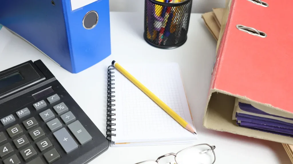 An office desk featuring a calculator, ring binders, a notebook, and eyeglasses for administrative work.