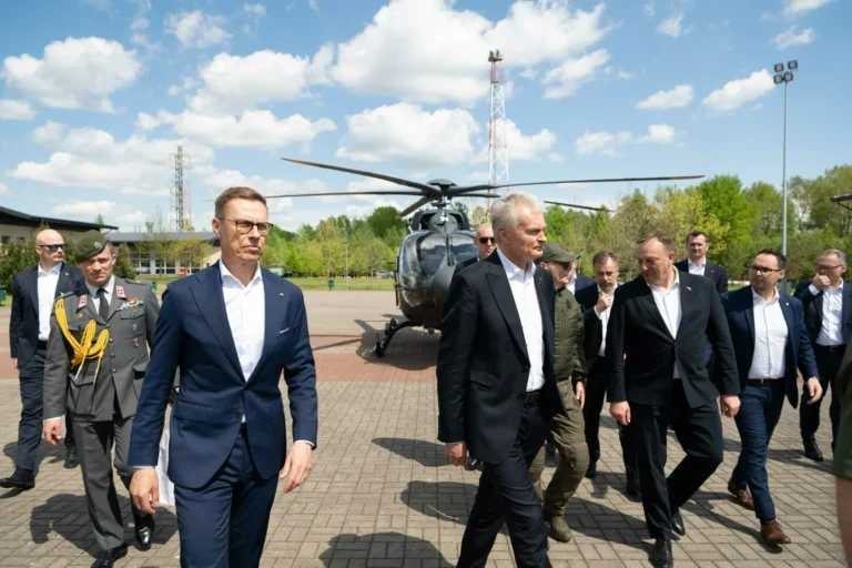 Two men in suits walk on pavement with a helicopter and military personnel in the background.