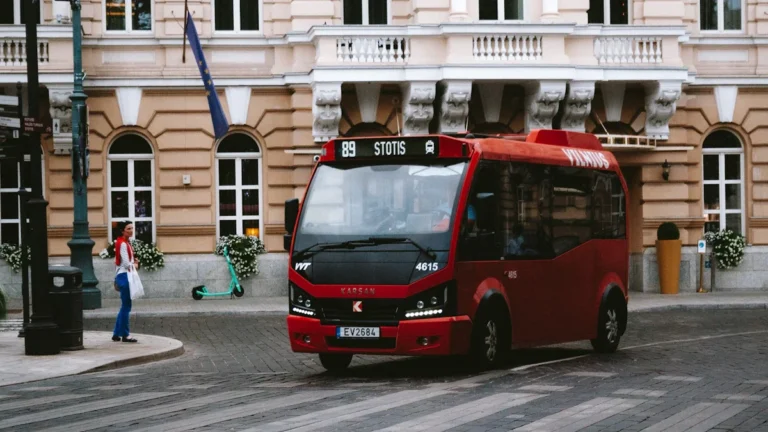 A modern red public bus with 'STOTIS' on its display driving through a street in Vilnius, Lithuania.