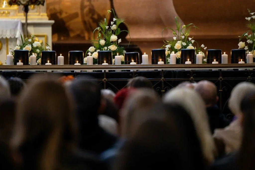 Row of black urns surrounded by white roses and candles at a memorial service with mourners.