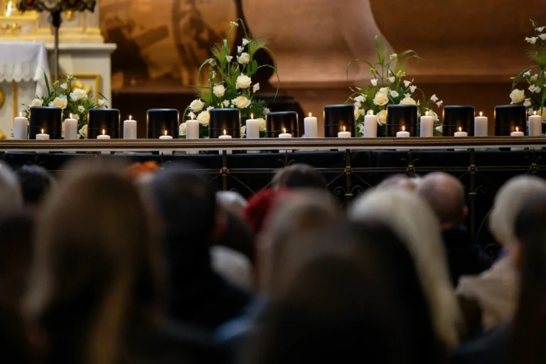 Row of black urns surrounded by white roses and candles at a memorial service with mourners.