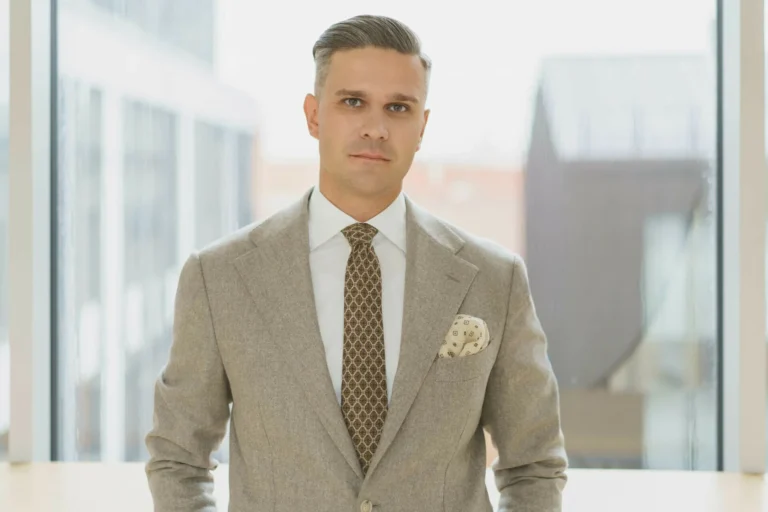 A man in a beige suit and patterned tie stands against a bright, blurred office window background.