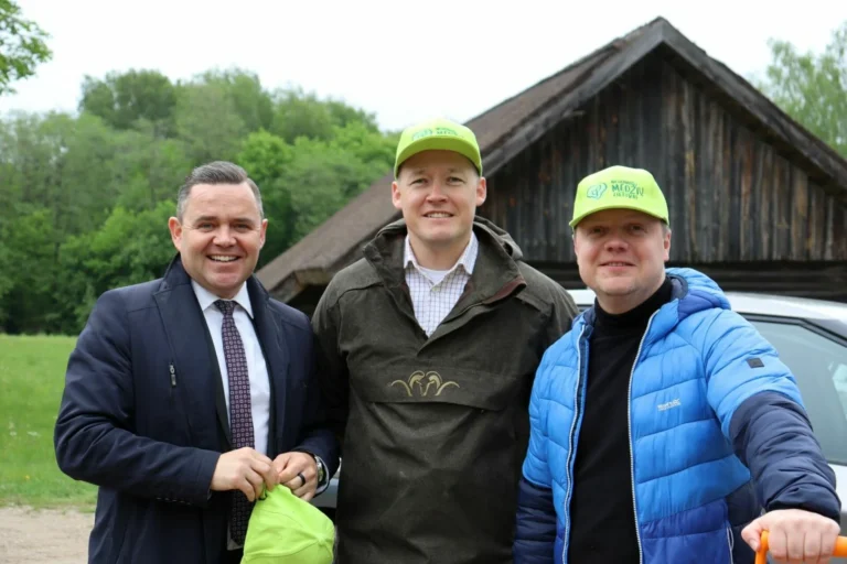 Three men smile outdoors in front of a rustic wooden barn; two wear neon green caps.