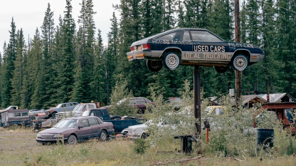 A weathered used car mounted on a tall pole above a rural scrapyard with evergreen trees.