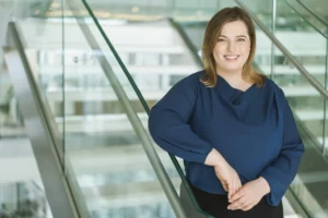 Professional woman in a blue blouse smiling while leaning against a modern glass railing.
