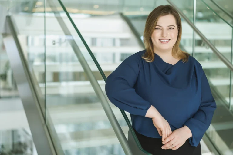 Professional woman in a blue blouse smiling while leaning against a modern glass railing.