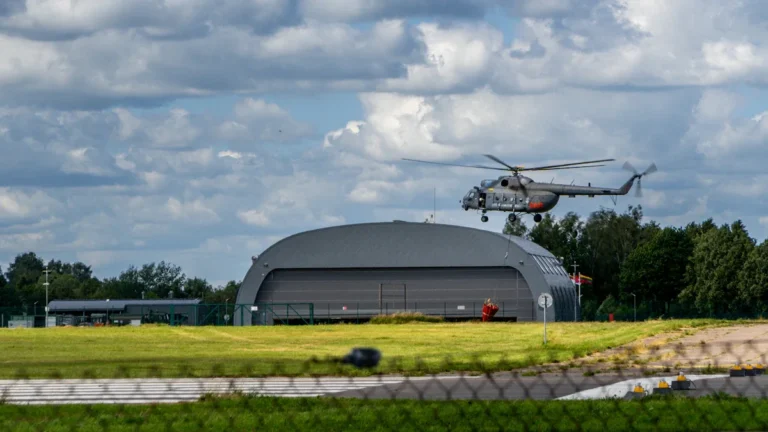 A military helicopter maneuvers over an airfield hangar in Lithuania, representing the nation's defense infrastructure.