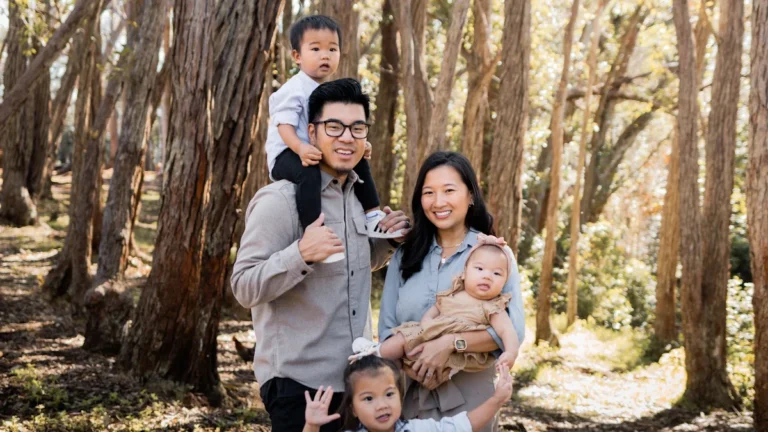 A smiling family of five with three children stands together in a sun-streaked woodland setting.