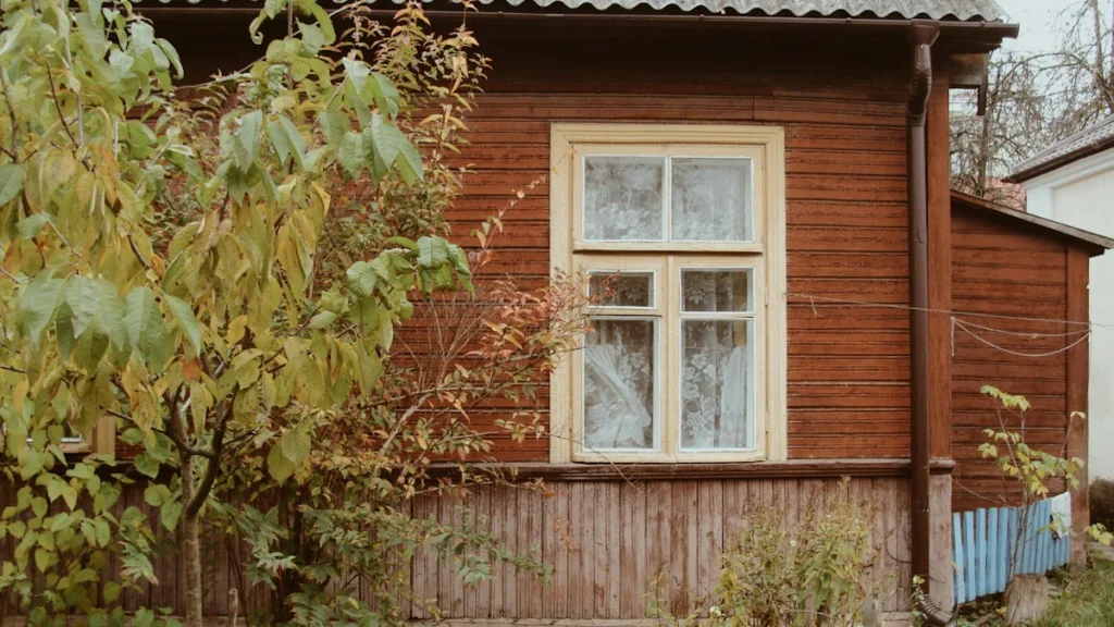 A traditional brown wooden garden house with a white window frame surrounded by lush green foliage.