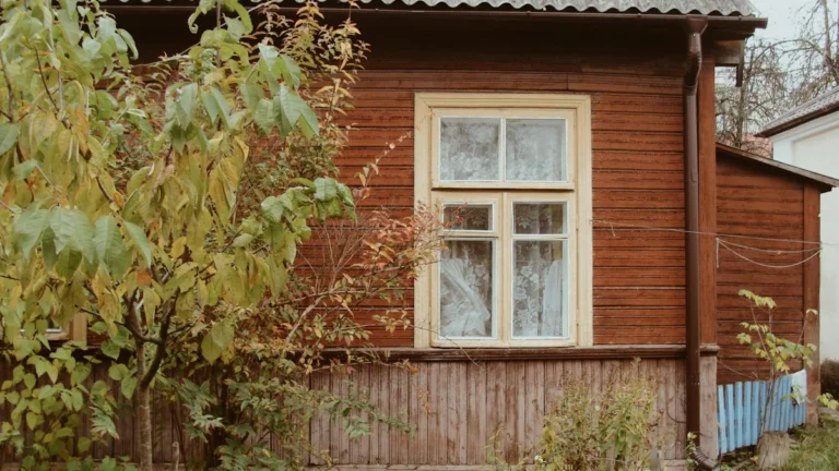 A traditional brown wooden garden house with a white window frame surrounded by lush green foliage.