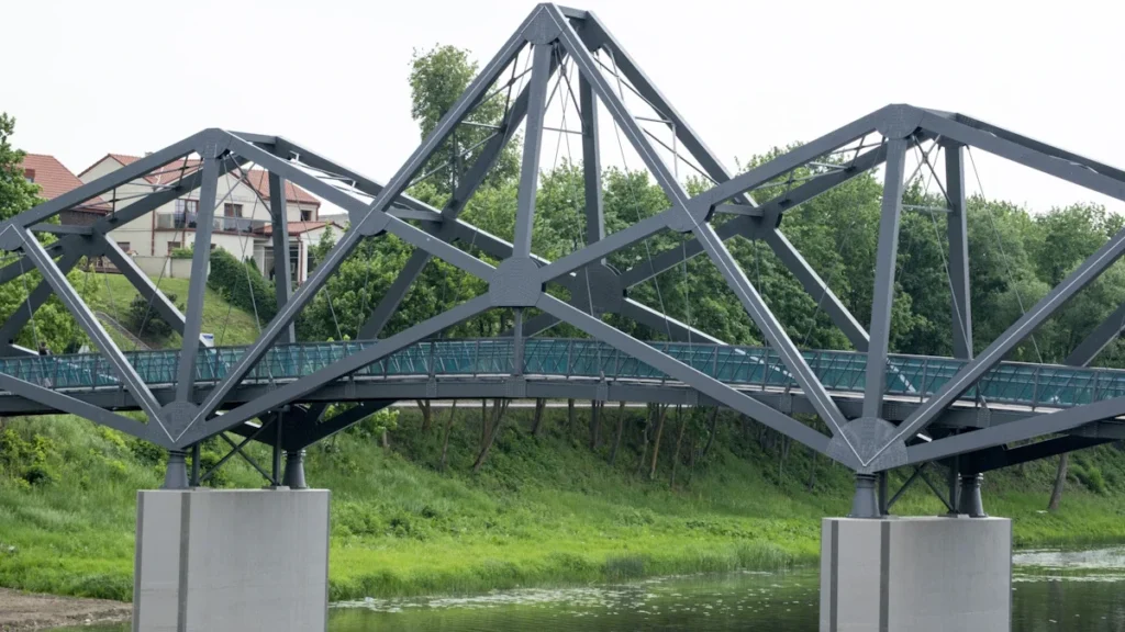 A modern grey metal truss bridge spanning a river with a building in the background in Lithuania.