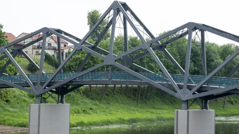 A modern grey metal truss bridge spanning a river with a building in the background in Lithuania.