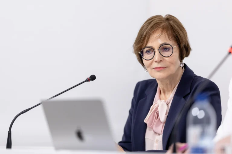 Professional woman with glasses wearing a navy blazer seated behind a microphone and laptop.