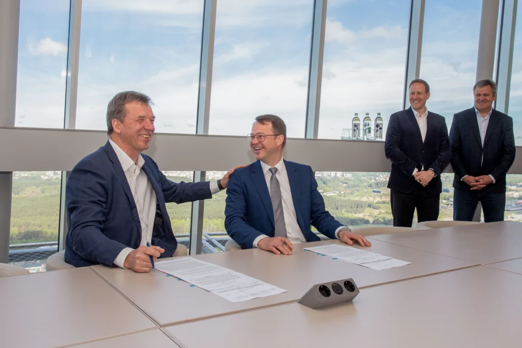 Two businessmen sit at a table signing documents and laughing in a high-rise office.