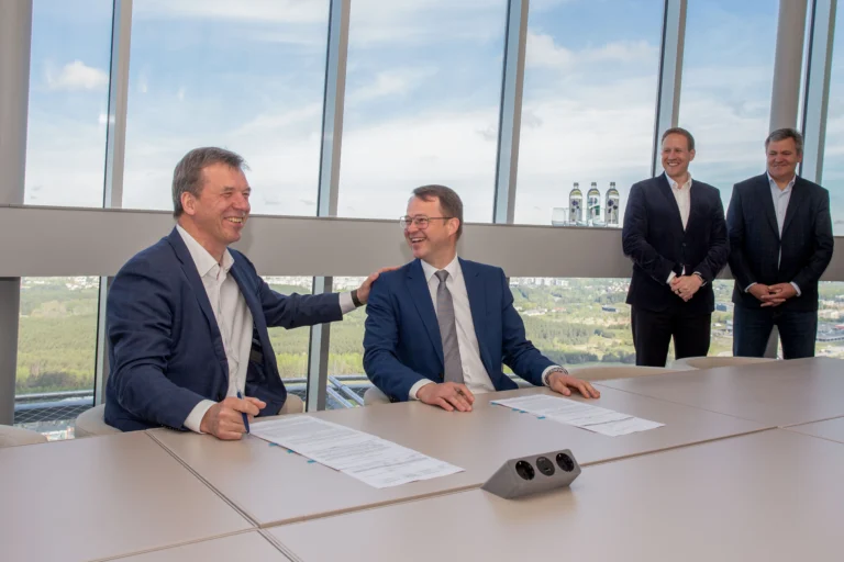 Two businessmen sit at a table signing documents and laughing in a high-rise office.