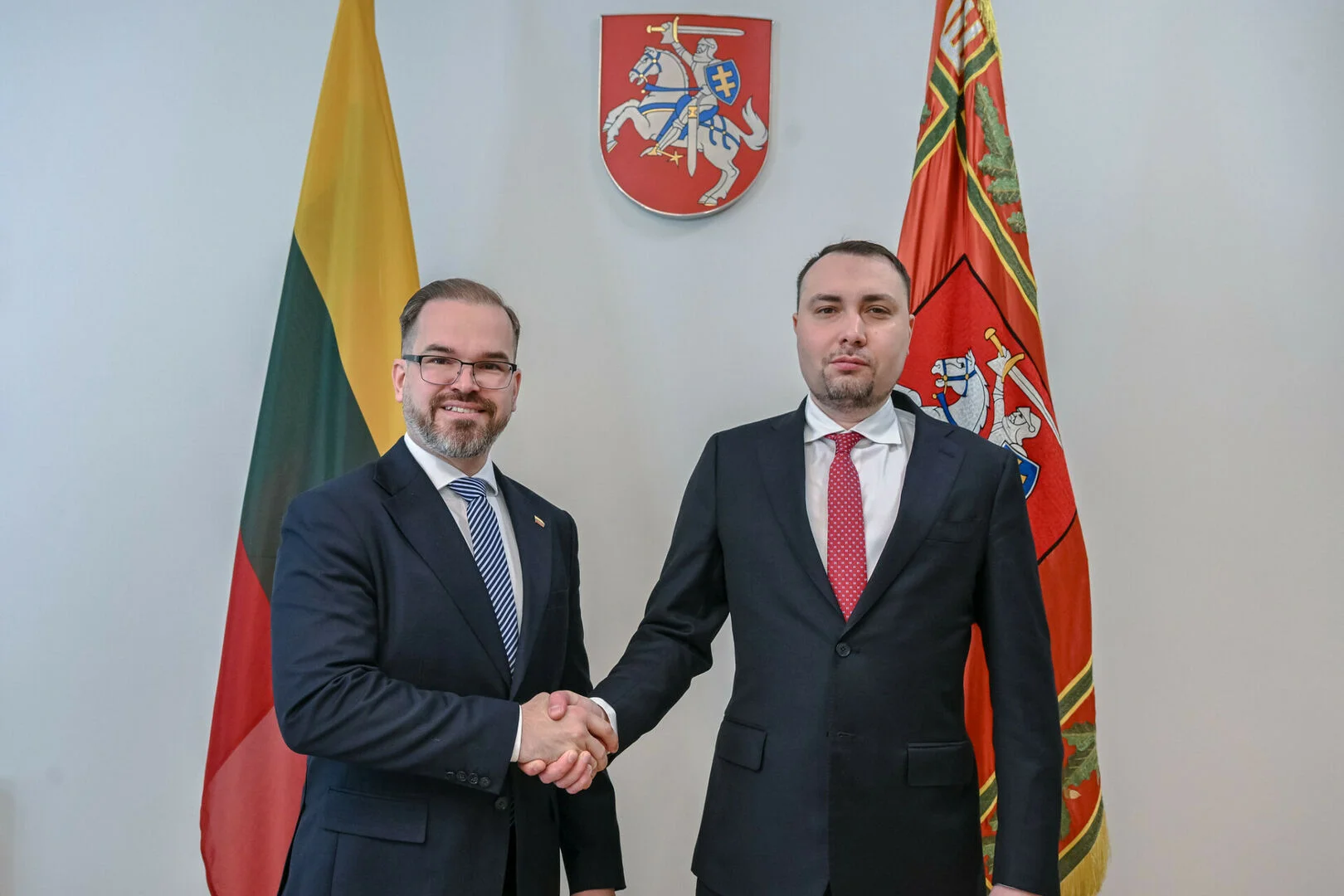 Two men in suits shaking hands in front of Lithuanian flags and a national coat of arms.
