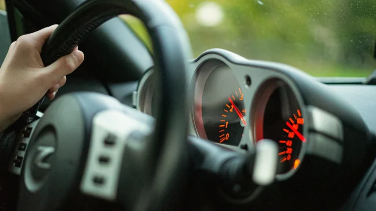 A person's hand on a car steering wheel with the dashboard gauges visible in soft focus.