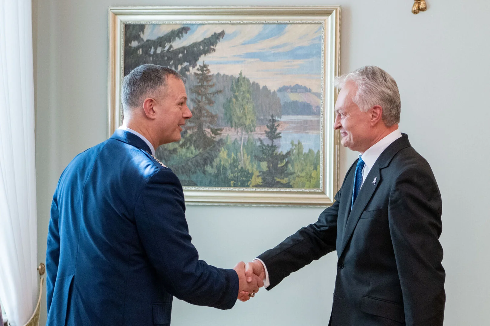 Military officer in blue uniform shakes hands with a man in a dark suit indoors.