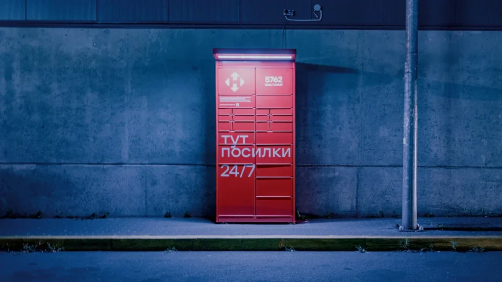 A bright red 24/7 parcel locker stands against a concrete wall at night, representing the infrastructure being utilized for civil defense in Lithuania.