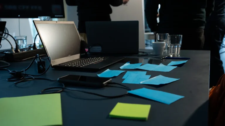 A laptop and several blue sticky notes on a desk in a busy modern office setting.