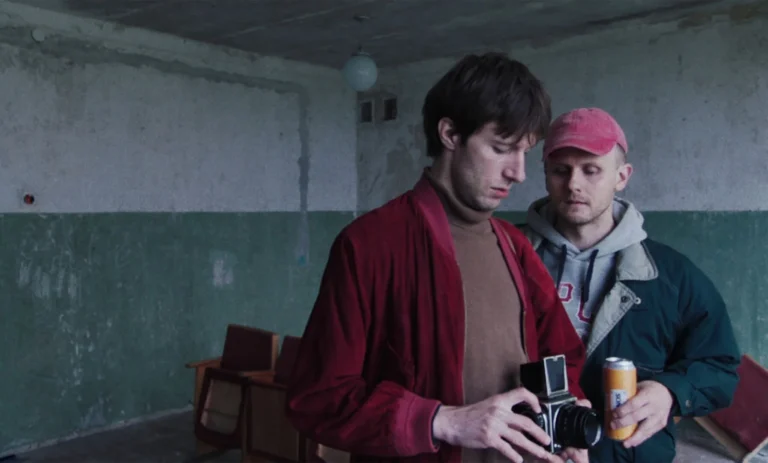 Two men in a dim room; one holds a vintage camera, the other a beverage.