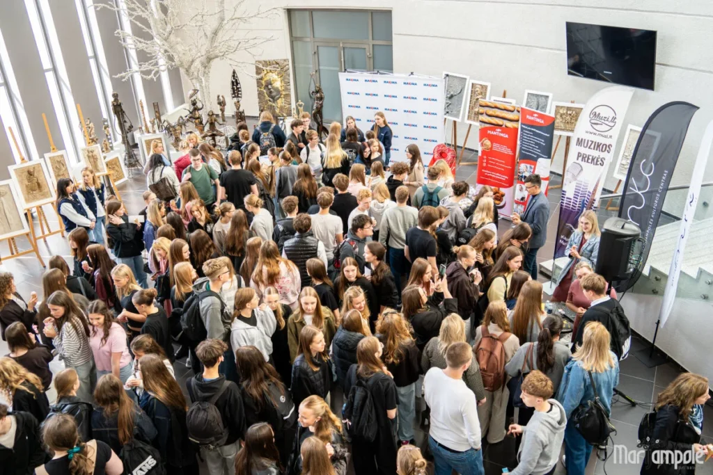 A large crowd of students gathers in a spacious hall featuring informational banners and displayed artwork.