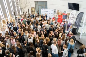 A large crowd of students gathers in a spacious hall featuring informational banners and displayed artwork.