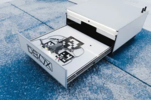 Small gray drone resting on a landing pad inside an open white DBOX storage unit.