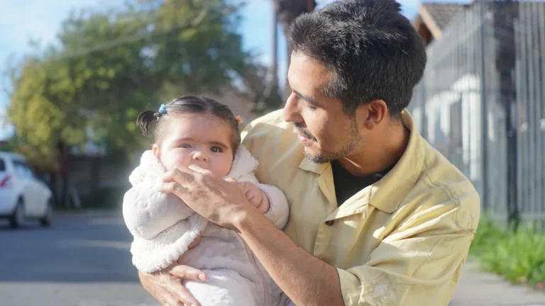 A father in a yellow shirt holds his young baby daughter outdoors in the sunlight.