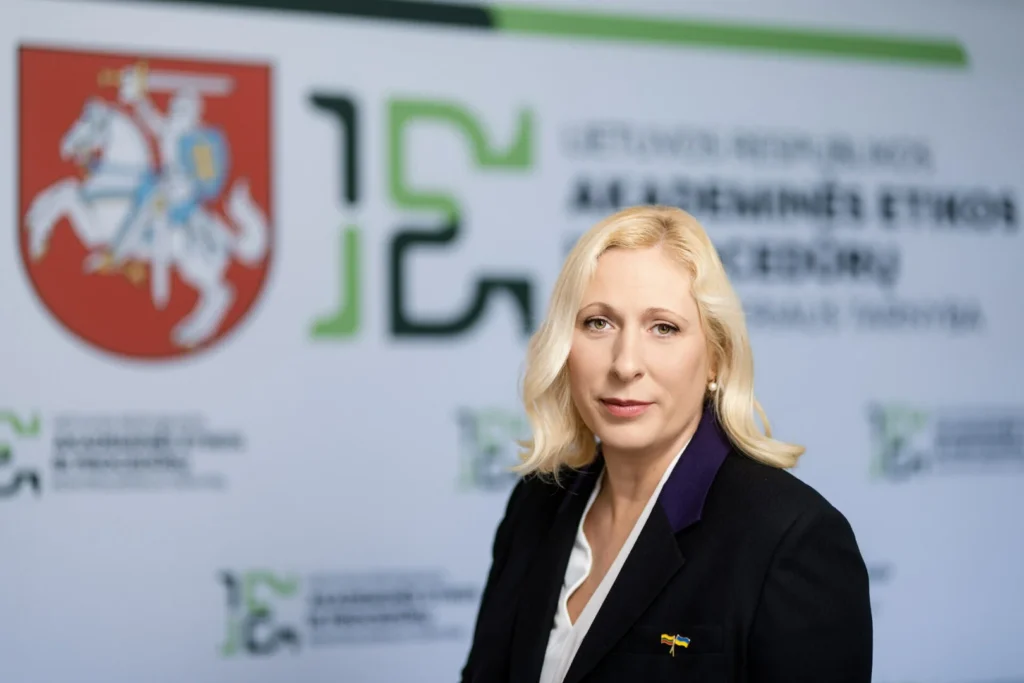 A blonde woman in a black blazer stands before a wall featuring the Lithuanian coat of arms.