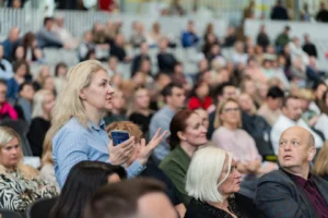 Blonde woman in blue shirt speaking and gesturing with a smartphone in a crowded public hall.