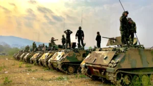 A line of military armored personnel carriers with soldiers standing on top against a cloudy sky.