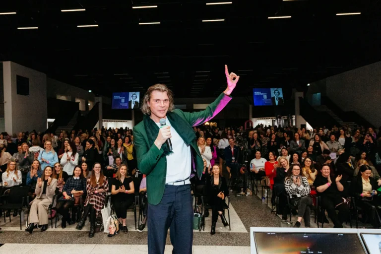 A man in a green jacket speaks with a microphone before a large seated audience indoors.