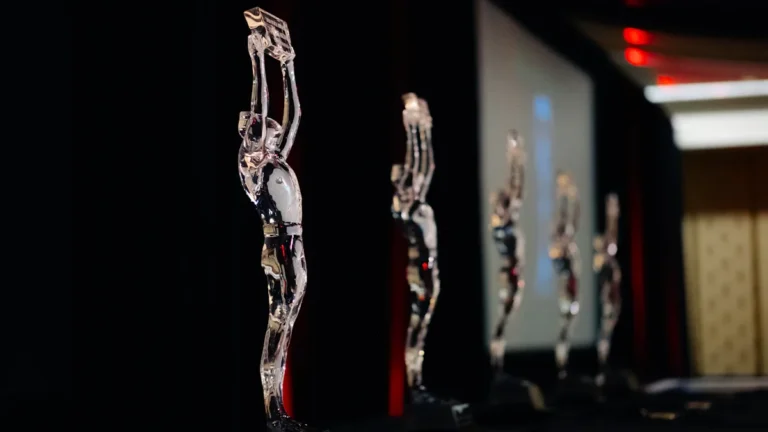 Several clear glass award statuettes shaped like human figures stand in a row under event lighting.