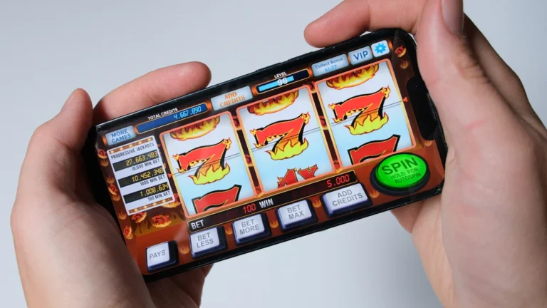 A person holds a smartphone displaying a colorful digital slot machine game with spinning reels.