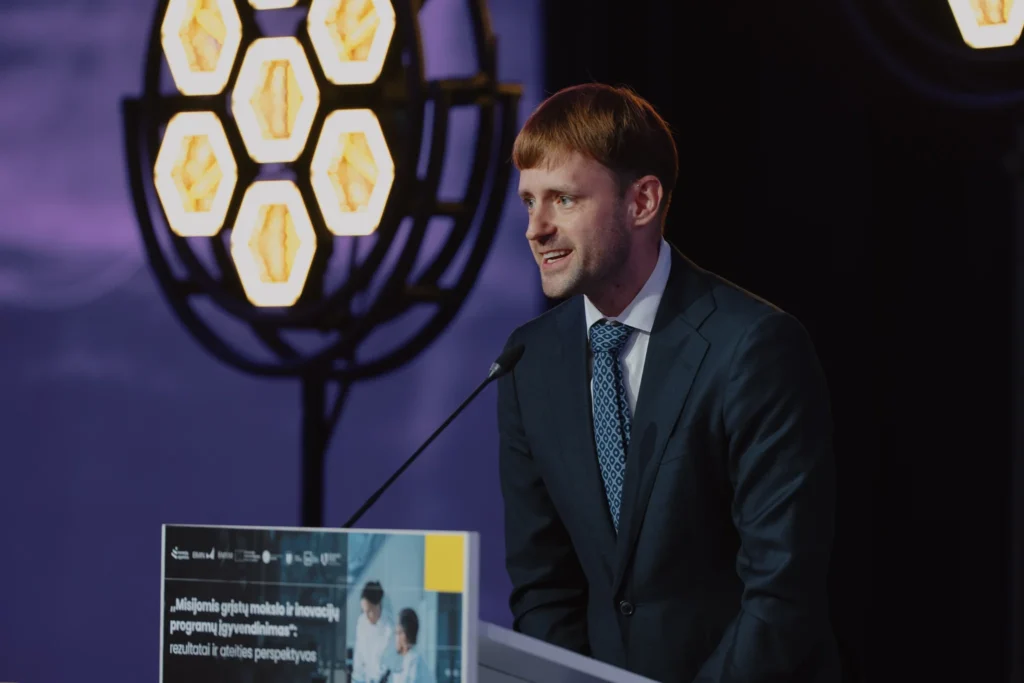 A man in a suit and patterned tie speaking at a podium with hexagonal stage lights.