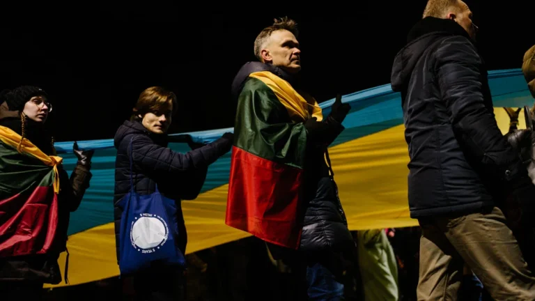 Lithuanian citizens draped in the national flag gather during a public demonstration at night.