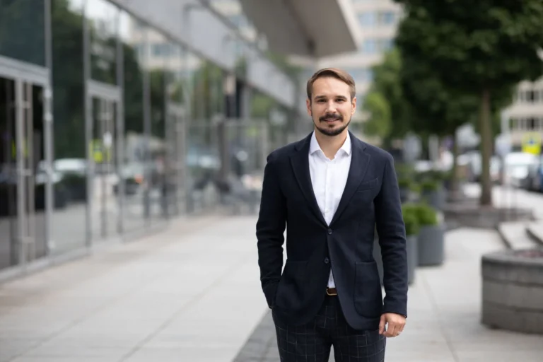 Smiling bearded man in a blazer and white shirt standing outside a modern office building.