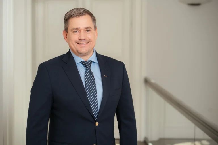 A smiling man in a navy blue suit and striped tie poses for a professional portrait.