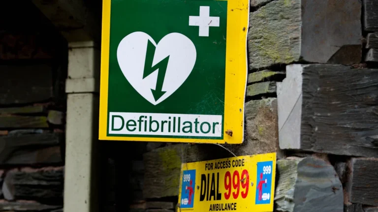 A green and yellow defibrillator sign mounted on a stone wall with emergency instructions to dial 999.
