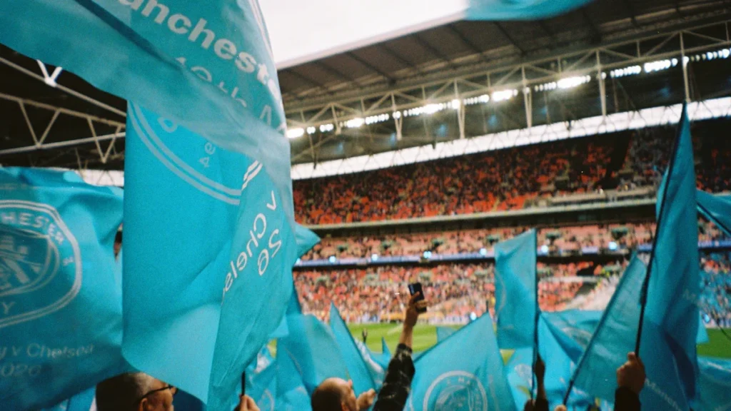 Manchester City football fans waving light blue flags in a large crowded stadium.