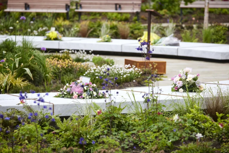 Colorful floral tributes resting on white marble memorial slabs in a lush, green landscaped garden.