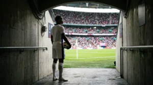 Football player in white kit stands in a tunnel looking out at a crowded stadium pitch.