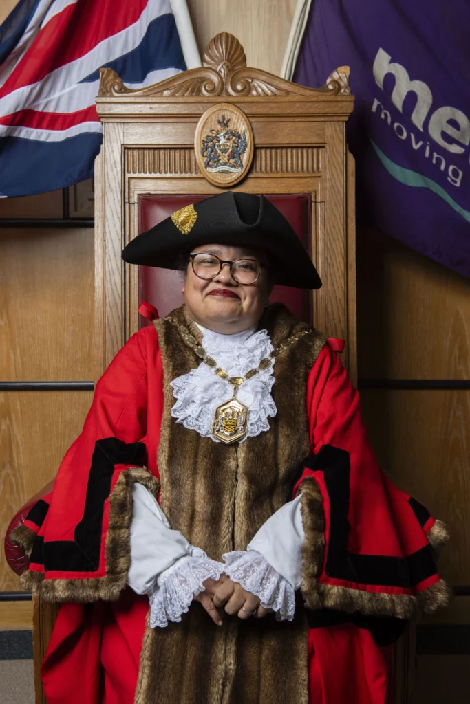 Official wearing red ceremonial robes and black bicorne hat sitting in a carved wooden chair.