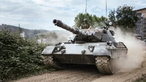 A modern military battle tank maneuvers through a dusty field during a daytime training exercise.
