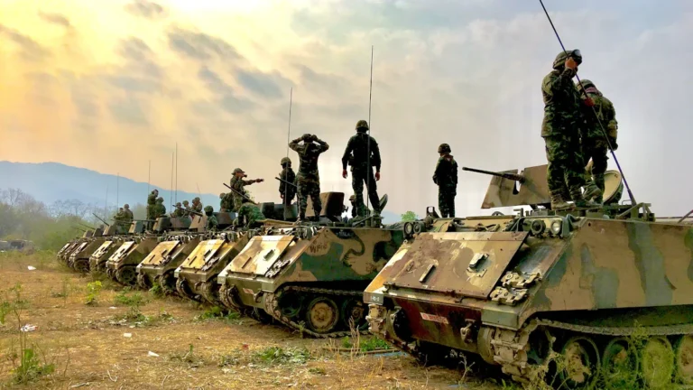 A line of military armored personnel carriers with soldiers standing on top during a training exercise.