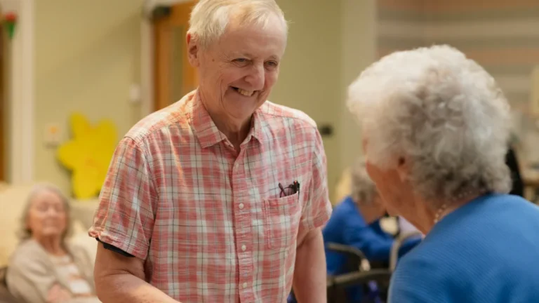 A smiling senior man in a checkered shirt talking with another person in a community common room.
