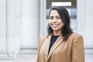 Smiling woman with dark hair in a tan blazer standing before large stone columns.